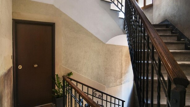 Una escalera con barandilla de madera y balaustradas de metal, que lleva a un rellano con una puerta y una planta en maceta.