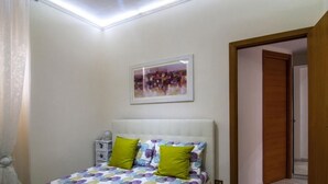 Un dormitorio con cama, almohadas, una mesita de noche, un cuadro en la pared y una puerta que da a otra habitación.