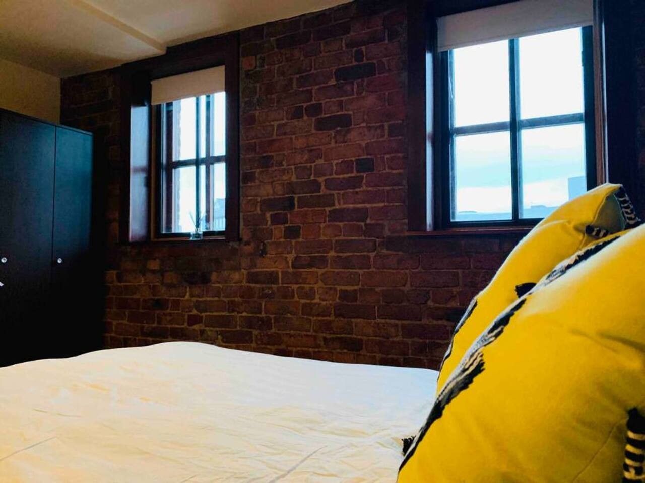 A room with a brick wall, a window, a bed with a white mattress, and a yellow pillow.
