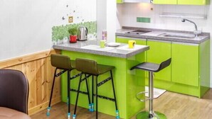 Una cocina compacta con un banco y taburetes de madera, una cama litera con barandillas verdes y una pequeña isla con un lavabo y taburetes.