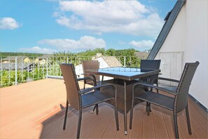 Um deck de madeira com mesa e cadeiras, guarda-corpo de metal e vista para casas ao fundo.