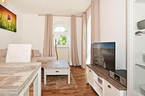 Ein Zimmer mit einer Couch, einem kleinen Tisch, einem Fernseher und einem Fenster mit Vorhängen.