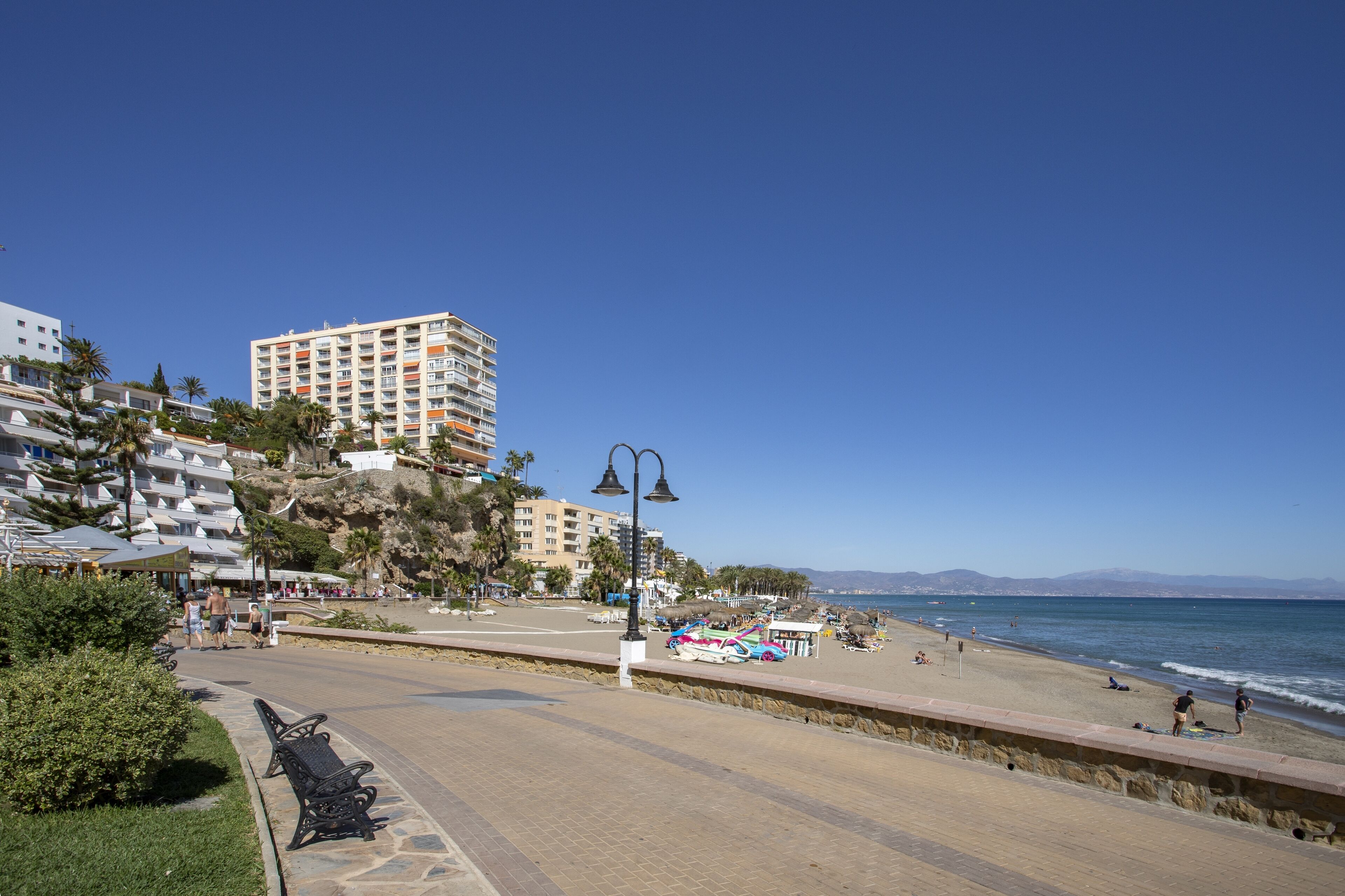 Un paseo marítimo con playa, un hotel de gran altura y cielo azul despejado.