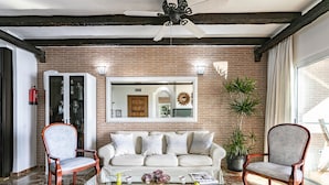 Ein Wohnzimmer mit Backsteinwand, einem weißen Sofa, zwei Holzstühlen, einem Glastisch und einem Deckenventilator.