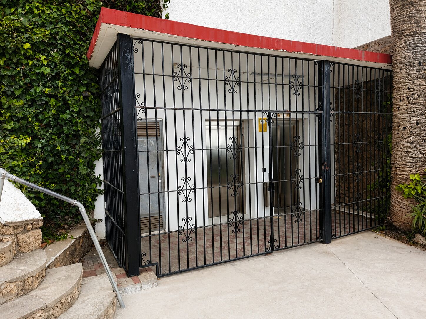 Una entrada protegida por un portón metálico, una escalera y un edificio con techo rojo.