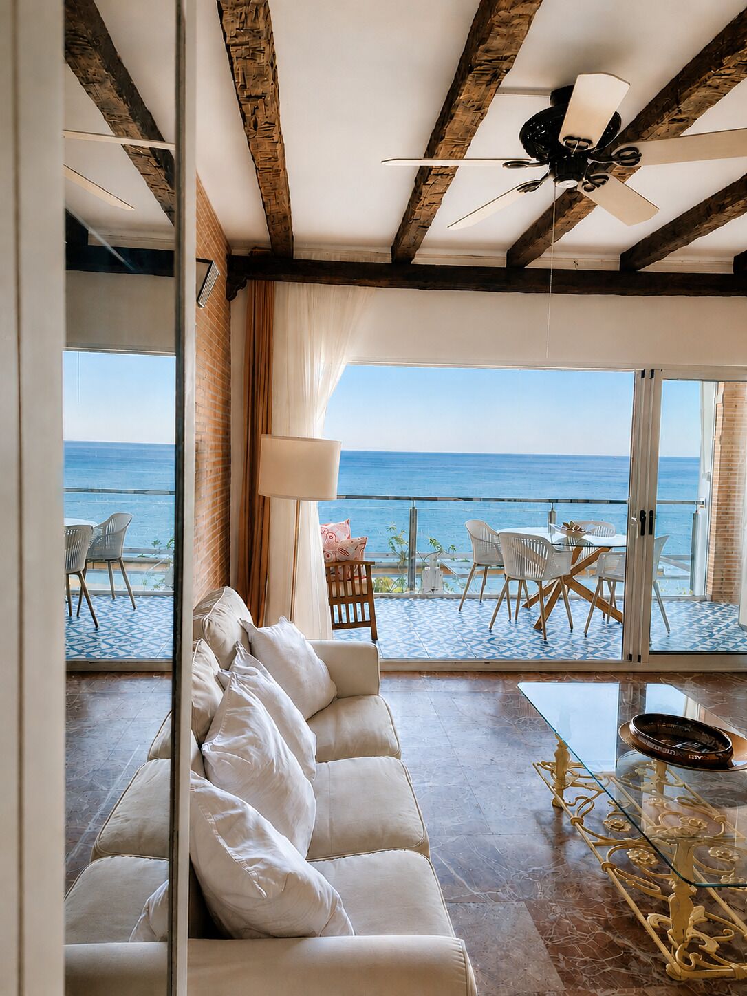 Una sala de estar moderna con un sofá blanco, una mesa de centro de vidrio y un ventilador de techo. Un amplio ventanal ofrece vistas al océano y a un patio con mobiliario exterior.
