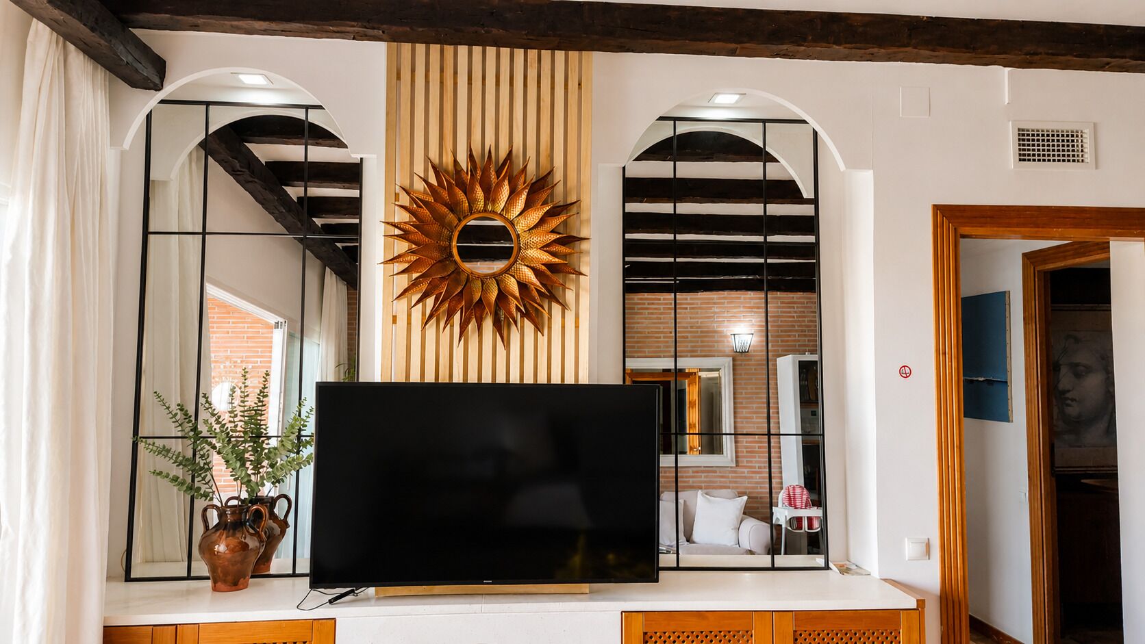A living room with a large flat-screen TV, a decorative sunburst mirror, and a potted plant.