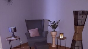 A living room with a grey armchair, a small table with a lamp, a potted plant, a floor lamp, and a decorative wall piece.