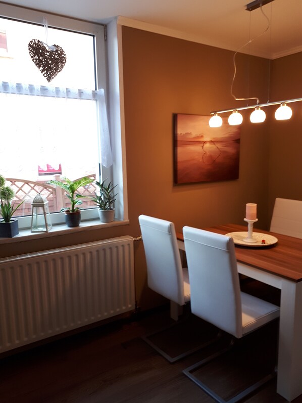 A dining area with a wooden table, white chairs, a potted plant, a radiator, and a window with a heart-shaped decoration.