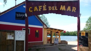 A café named Café del Mar with a wooden sign, a blue and red building, and a view of a lake.