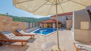 A pool area with lounge chairs and an umbrella, surrounded by stone walls and a view of green hills. - Stunning home in Krivodol (Krivodol)