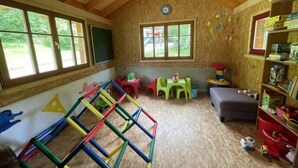 Een kamer met een houten plafond, grote ramen, een speelhoek met kleurrijk materiaal en een boekenplank.