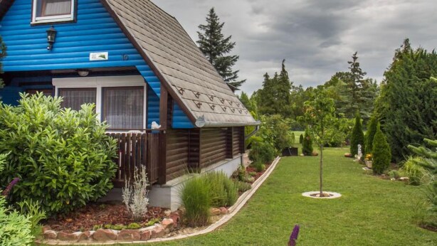 Une cabane en bois bleue, avec un toit en pente, un petit porche et un jardin bien entretenu où poussent différentes plantes, ainsi qu’une pelouse.