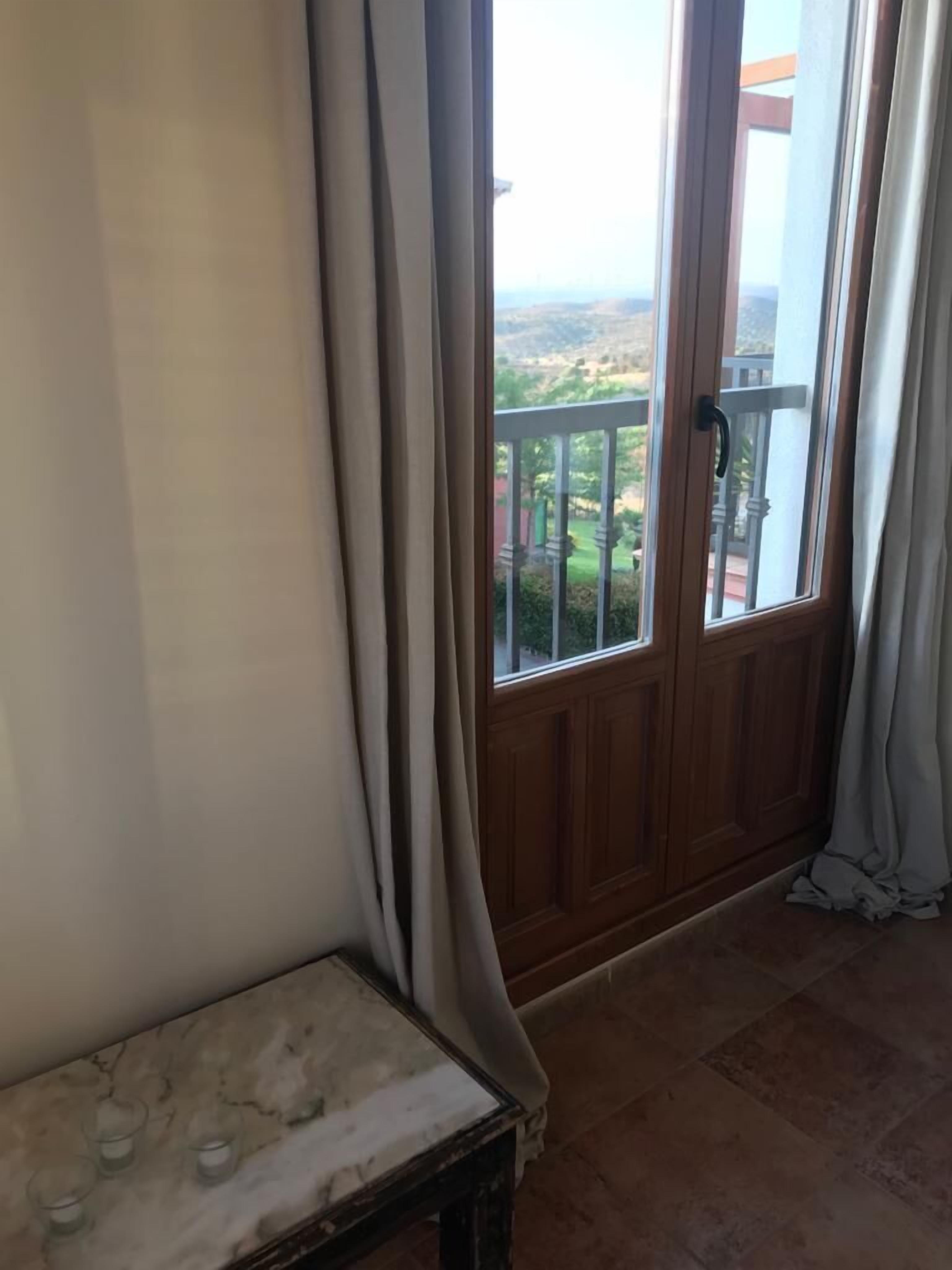 Ein Zimmer mit Holztür, die auf einen Balkon führt, einem kleinen Tisch mit Gläsern und Blick auf eine grüne Landschaft.