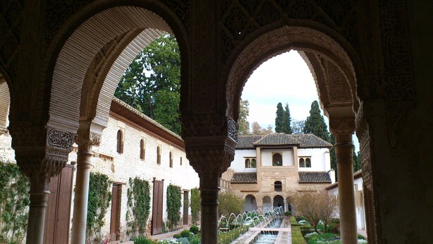 Un passage architectural bordé d’arcades, une fontaine et un bâtiment doté de fenêtres en arc de cercle et d’un toit recouvert de tuiles.