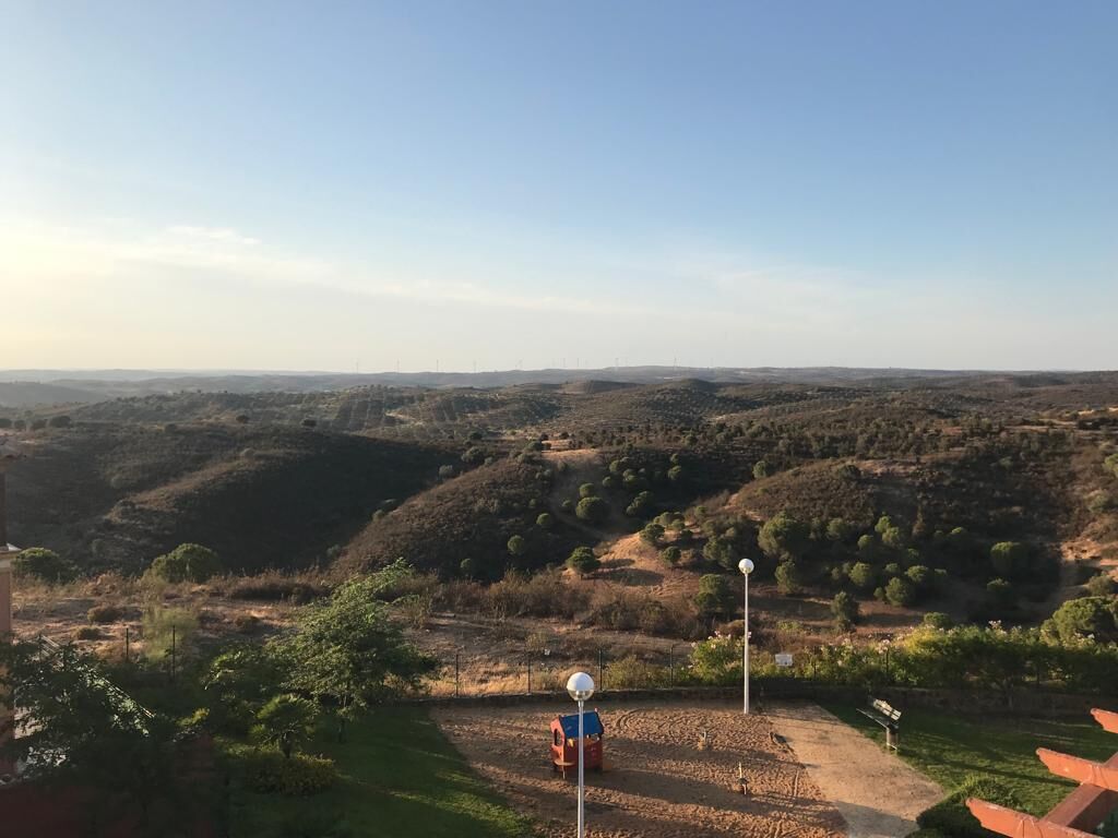 Een panoramisch uitzicht op een glooiend landschap, met een klein gebouw en een rood voertuig op de voorgrond.