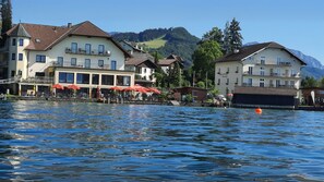 Ein Hotel am Seeufer mit mehreren Balkonen, Außenbereich zum Sitzen und Blick auf See und Berge.