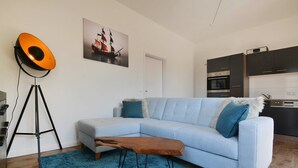 A modern living room with a light blue sofa, a small wooden bench, a blue rug, a floor lamp, and a kitchen in the background. - Cozy vacation apartment in Koserow with terrace - only 350 m to the beach (Koserow)