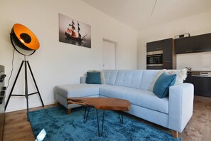 Un soggiorno moderno con un divano blu chiaro, una piccola panca in legno, un tappeto blu, una lampada da terra e una cucina sullo sfondo.