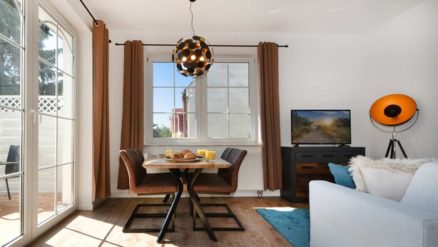 A modern living room with a dining area, a sofa, a TV, and a large window with curtains. - Cozy vacation apartment in Koserow with terrace - only 350 m to the beach (Koserow)