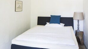 Een netjes opgemaakt bed met een donker hoofdbord, twee kussens, een bedlampje en een lijst met een foto aan de muur.