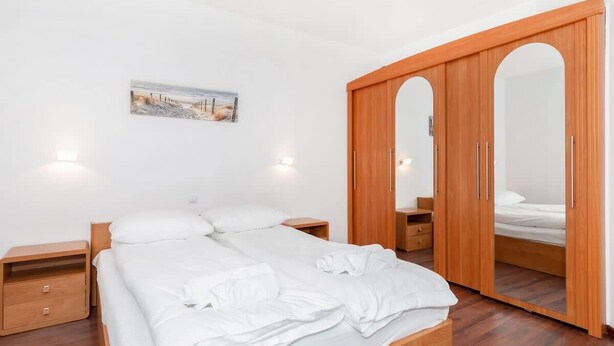 Una cama bien hecha con sábanas blancas, una mesita de noche de madera con cajón y un armario de madera con puertas espejadas, además de un cuadro en la pared.