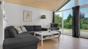 A modern living room with a sectional sofa, a white coffee table with books, a floor lamp, and a large window with a view of greenery. - Nice home in Jægerspris with kitchen (Jægerspris)
