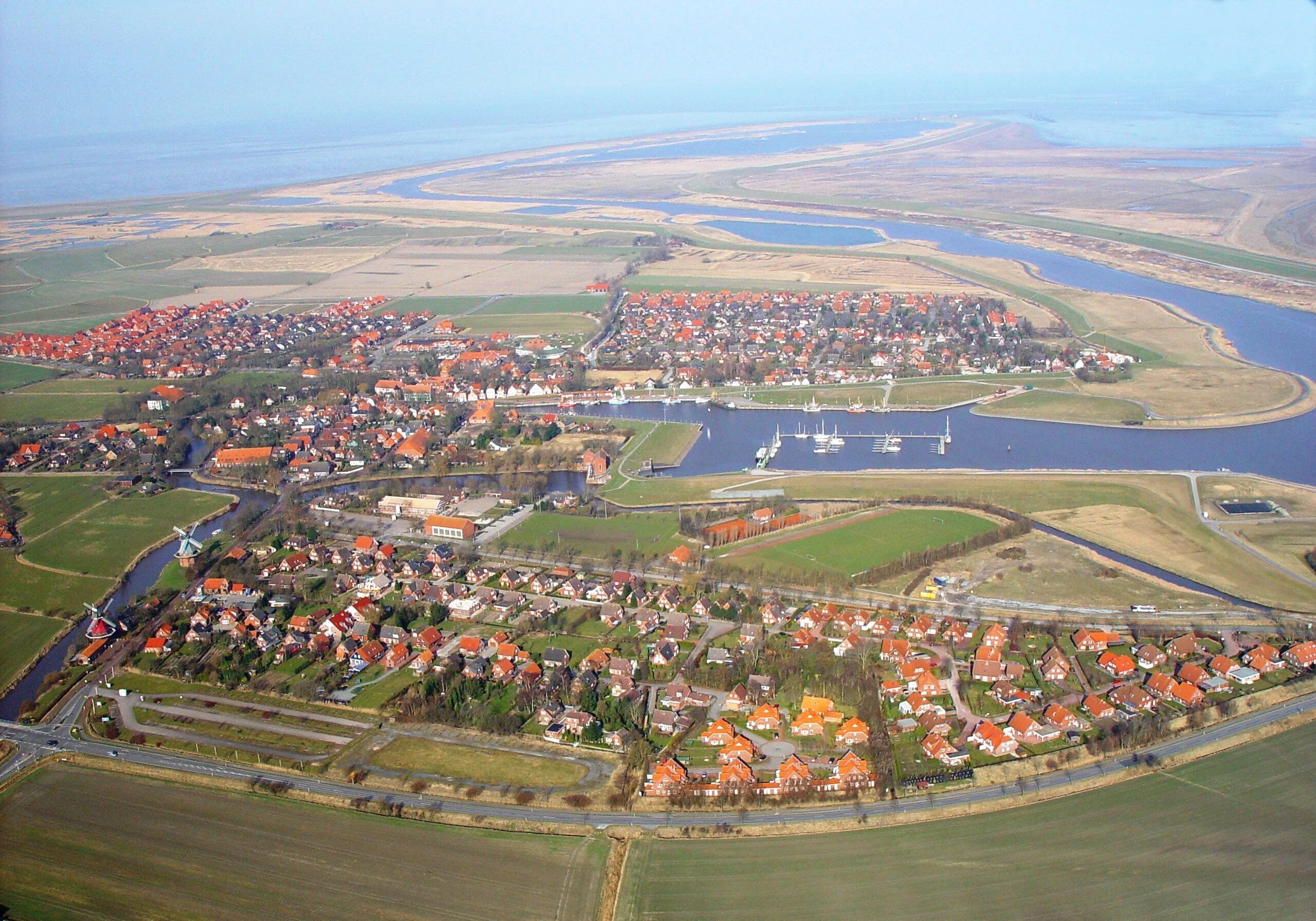Luchtbeeld van een kustplaatsje met rode daken, een jachthaven en een rivier.