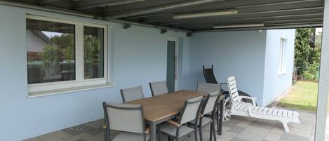 A covered patio with a table and chairs, a lounge chair, and a view of the outdoors.