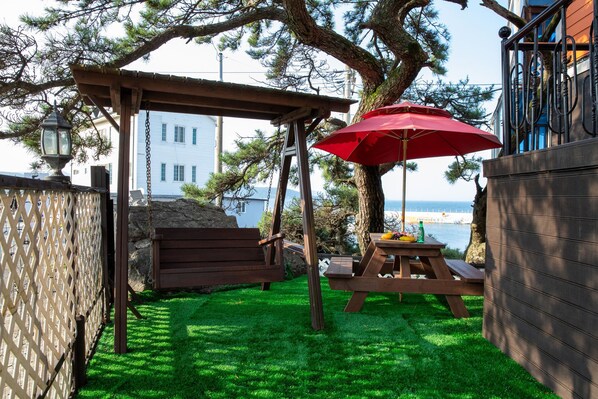 A wooden pergola with a red umbrella, a picnic table, and a bench in a backyard with a view of the ocean.