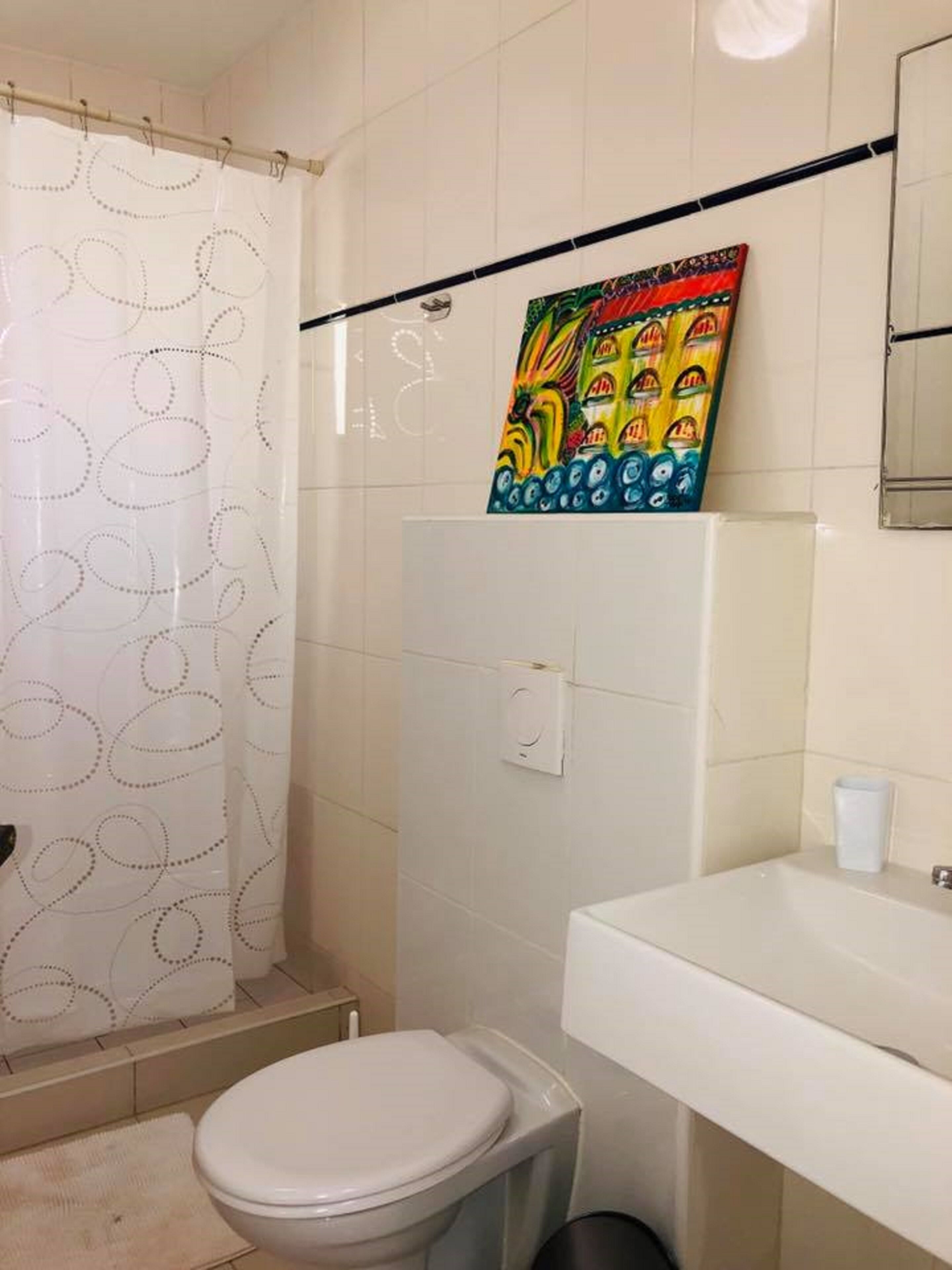 A bathroom with a toilet, a shower with a patterned curtain, a sink, and a colorful painting on the wall.