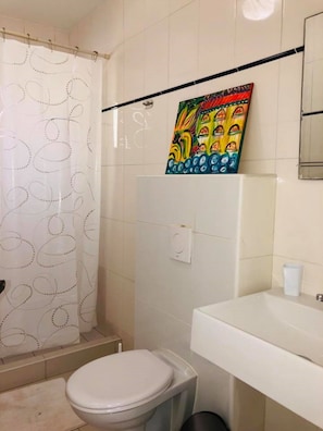 A bathroom with a toilet, a shower with a patterned curtain, a sink, and a colorful painting on the wall. - Villa Brakkeput 308 (Jan Thiel)
