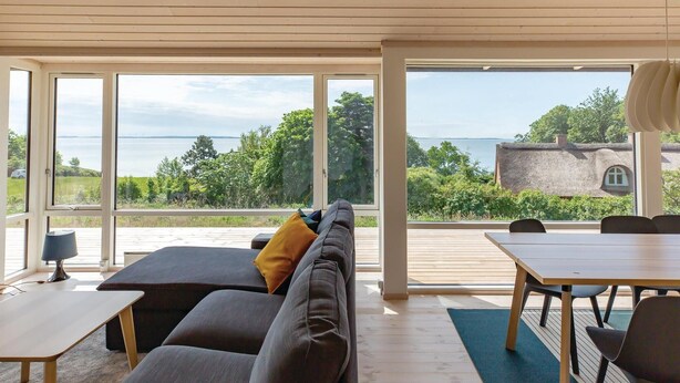 A modern living room with a sofa, a small table, and a dining table. Large windows offer a view of a lake and a thatched-roof building. - 3 bedroom nice home in Kalundborg (Kalundborg)