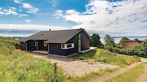 A modern house with a dark exterior, a sloped roof, and a large window. It is surrounded by greenery and a body of water in the background. - 3 bedroom nice home in Kalundborg (Kalundborg)