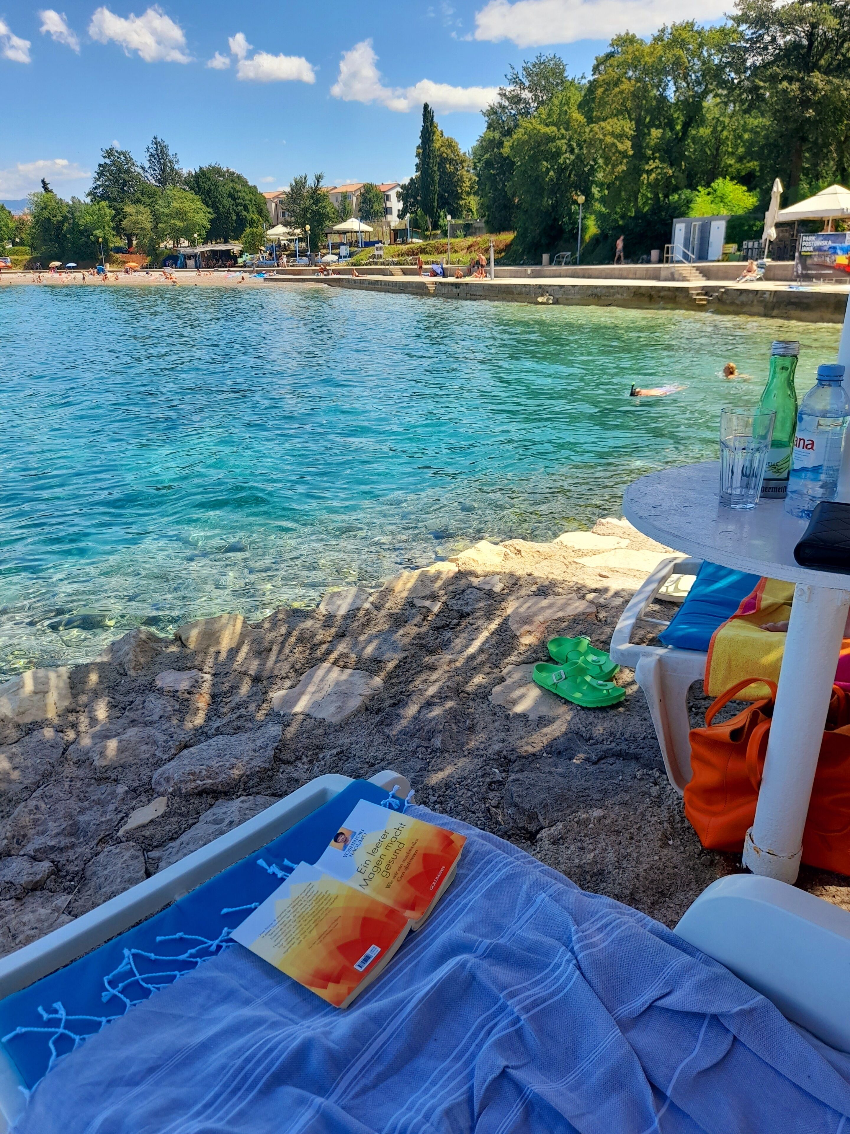 Un área junto a la piscina con agua cristalina, una silla de descanso, una mesa con un libro y una botella, y vistas a la piscina y los alrededores.