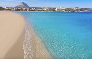 A beach with clear blue water, a sandy shore, and buildings in the background.