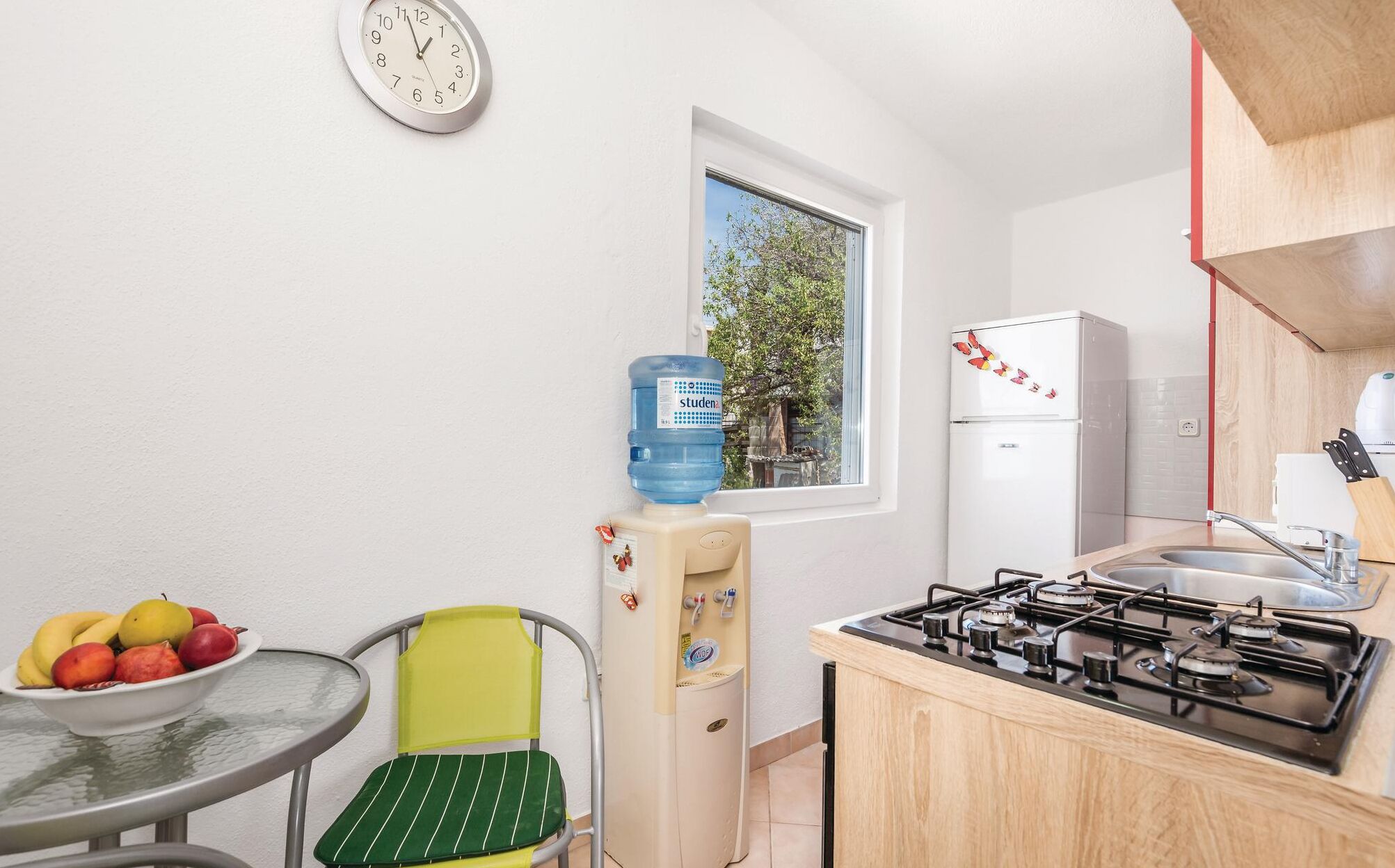 A compact kitchen with a gas stove, a water dispenser, a refrigerator, and a dining area with a chair and table.