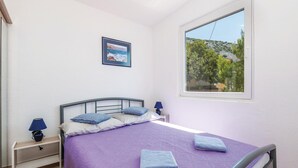 Un dormitorio con cama, mesita de noche, lámpara, un cuadro en la pared y ventana con vistas a zonas verdes.