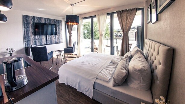 Habitación de hotel moderna con una cama grande, un escritorio con cafetera, un televisor montado en una pared texturizada y ventanas amplias con cortinas.