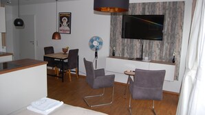Una sala de estar moderna con comedor, televisor montado en una pared de madera, un ventilador y una pequeña mesa con sillas.