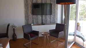 Una sala de estar moderna con un televisor de pantalla plana montado en una pared de madera, dos sillones oscuros, una mesita redonda, un jarrón con flores y un amplio ventanal con vistas al exterior.