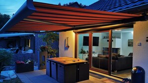 A modern outdoor patio with a wooden table and chairs, a glass door leading to a living area, and a brick wall.