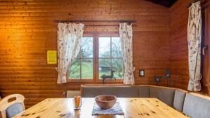 A wooden cabin interior with a dining table, chairs, a large window with curtains, and a view of the outdoors. - Reinskopf by Interhome (Schönecken)