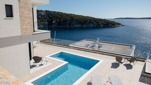 Un área de piscina moderna con vista al mar, mobiliario exterior y un muro de piedra.