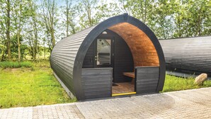 Een moderne, halfronde houten constructie met een glazen deur, omgeven door groen en een verhard pad.