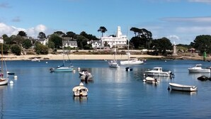 Un paysage côtier avec des bateaux amarrés dans des eaux calmes, un phare et des maisons le long de la côte.