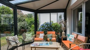 Une pièce baignée de lumière naturelle, dotée d’un plafond en verre et meublée de chaises et de canapés en bois, ainsi que d’une table basse et de diverses plantes.
