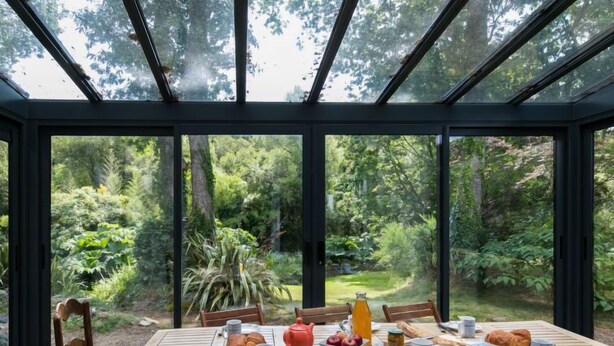 Un coin repas avec une table dressée pour le petit-déjeuner, entouré de grandes fenêtres donnant sur un jardin luxuriant.