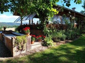 Une maison en bois avec un toit en pente, entourée d’un jardin où poussent différentes plantes et fleurs.