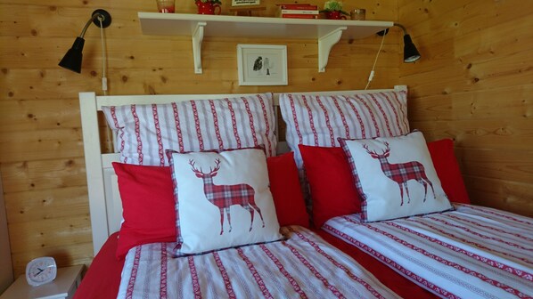Una habitación de madera con una cama, ropa de cama a rayas y almohadas decorativas, una mesita de noche con un reloj, y un estante con libros y un cuadro enmarcado.